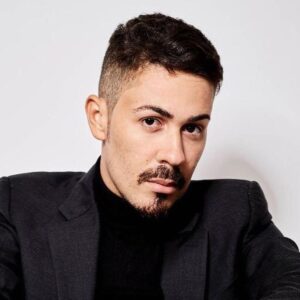 Carlinhos Maia posa para foto com blazer escuro e gola alta preta contra fundo branco.