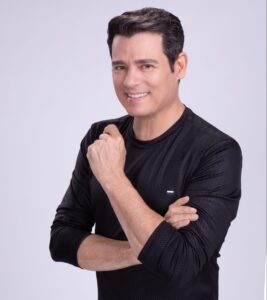 Celso Portiolli sorrindo, vestindo blusa preta de manga longa, com os braços cruzados contra fundo cinza claro.