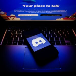 Smartphone com o logotipo do Discord sobre um notebook exibindo a página inicial da plataforma.