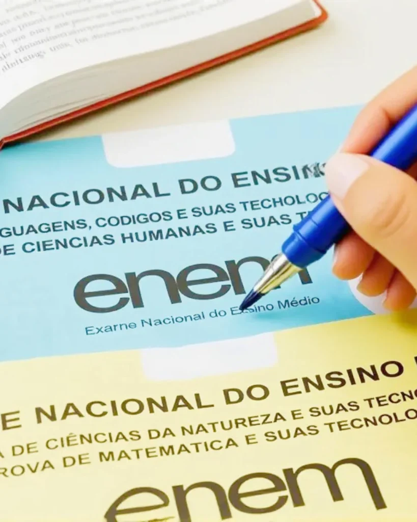 Mão segurando uma caneta azul sobre cadernos de prova do Enem.