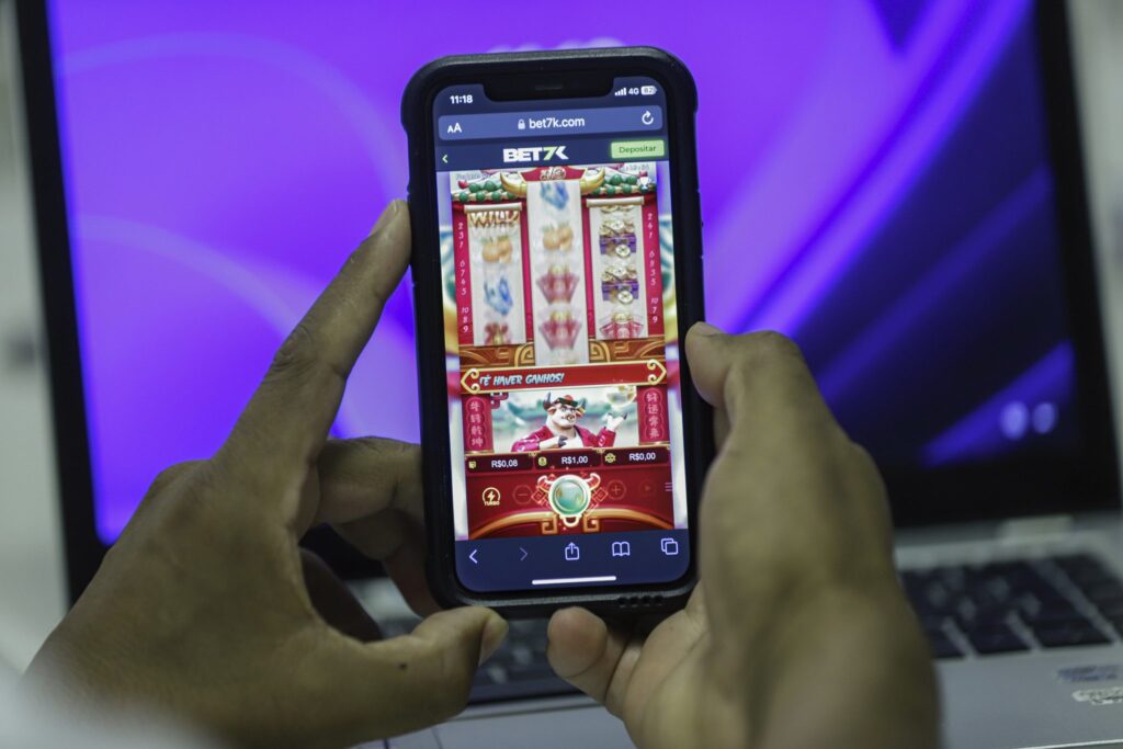 Mãos segurando um smartphone exibindo um jogo de apostas online em frente a um notebook.