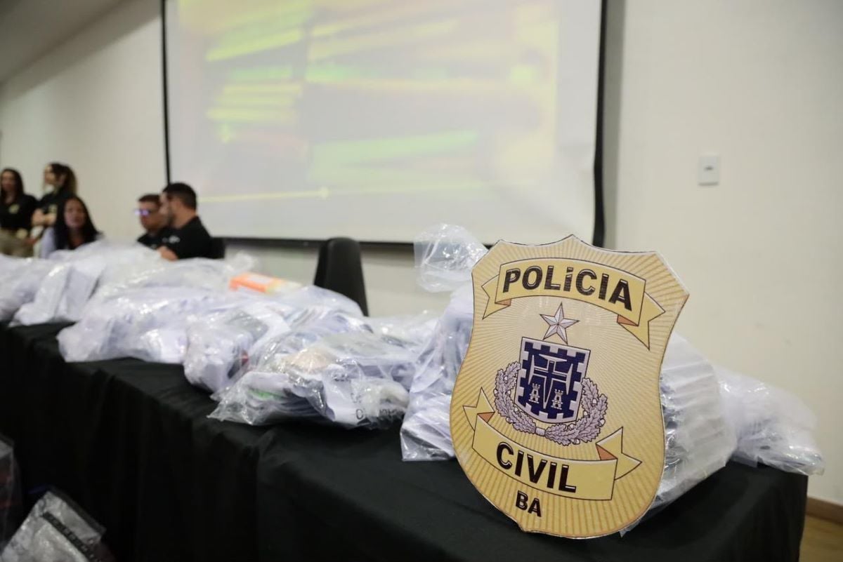 Distintivo da Polícia Civil da Bahia em destaque sobre mesa com celulares apreendidos em sacos plásticos.